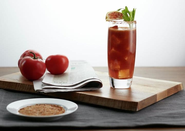 En la imagen se observa el cóctel Bloody Mary junto 3 tomates y un plato con comida.