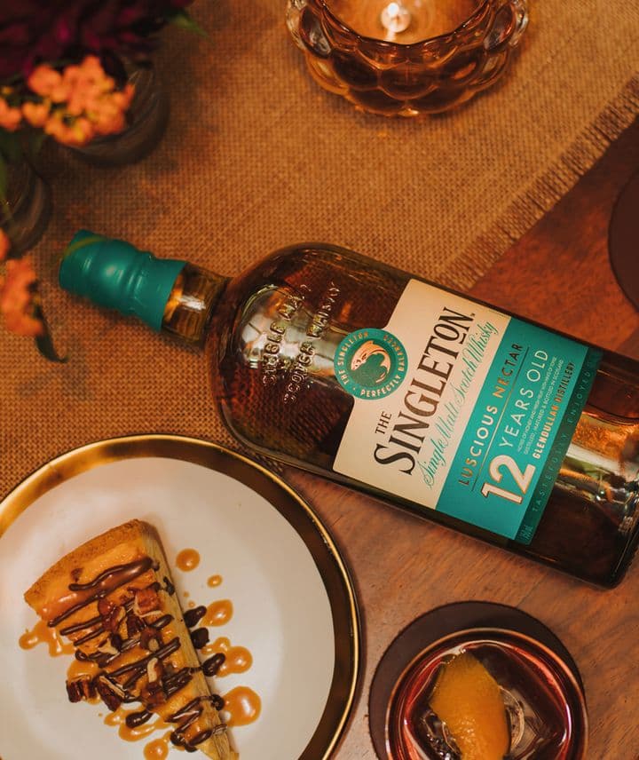 La imagen muestra una botella de whisky The Singleton 12 años junto a una porción de torta, y un vaso con el cóctel Old Fashioned.