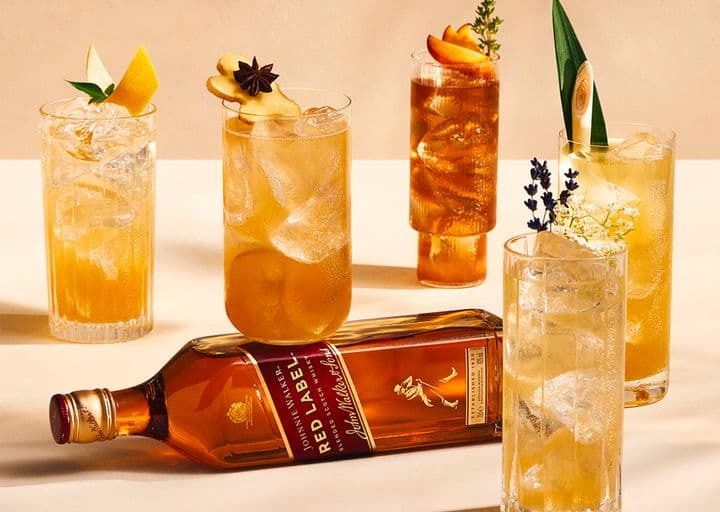 En la imagen se puede apreciar cinco vasos estilo highball de diferentes formas con una botella whisky Johnnie Walker Red Label apoyada de costado.