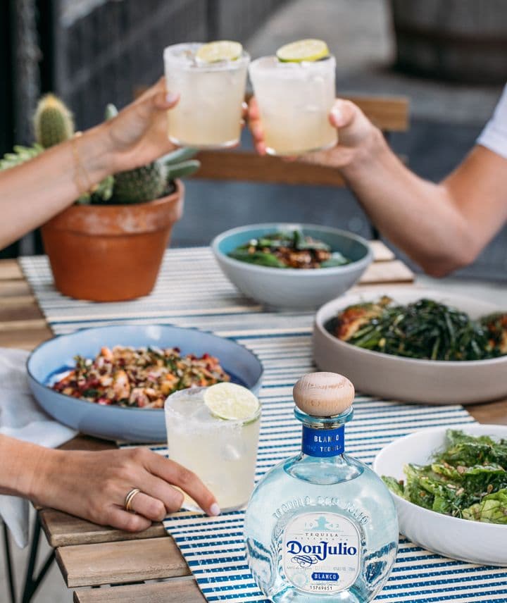 La imagen muestra una mesa con una variedad de platos de comida, destacando en el centro una botella de tequila Don Julio Blanco, mientras que dos personas brindan con cócteles Margarita.