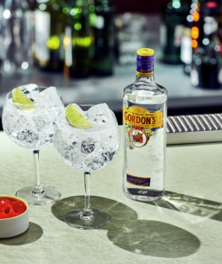Botella de gin Gordon´s, 2 copas de vidrio llenas de hielo, rodaja de limón en cada una como decoración y un pote con cerezas. Colocados en una barra