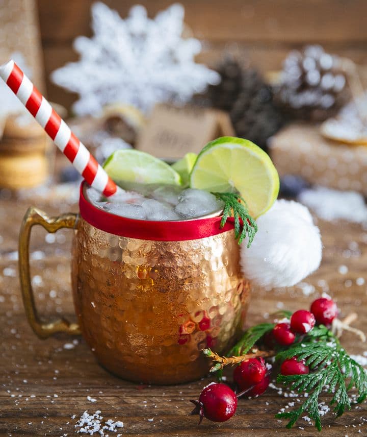 En la imagen se puede observar una taza de cobre con el cóctel "Moscow Mule" decorada con temática navideña, así como un entorno festivamente decorado acorde a la temporada navideña.
