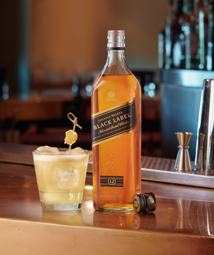 En la imagen se puede observar un vaso con el cóctel Penicillin y, al lado, una botella de Whisky Johnnie Walker Black Label, apoyados sobre una barra de bar.