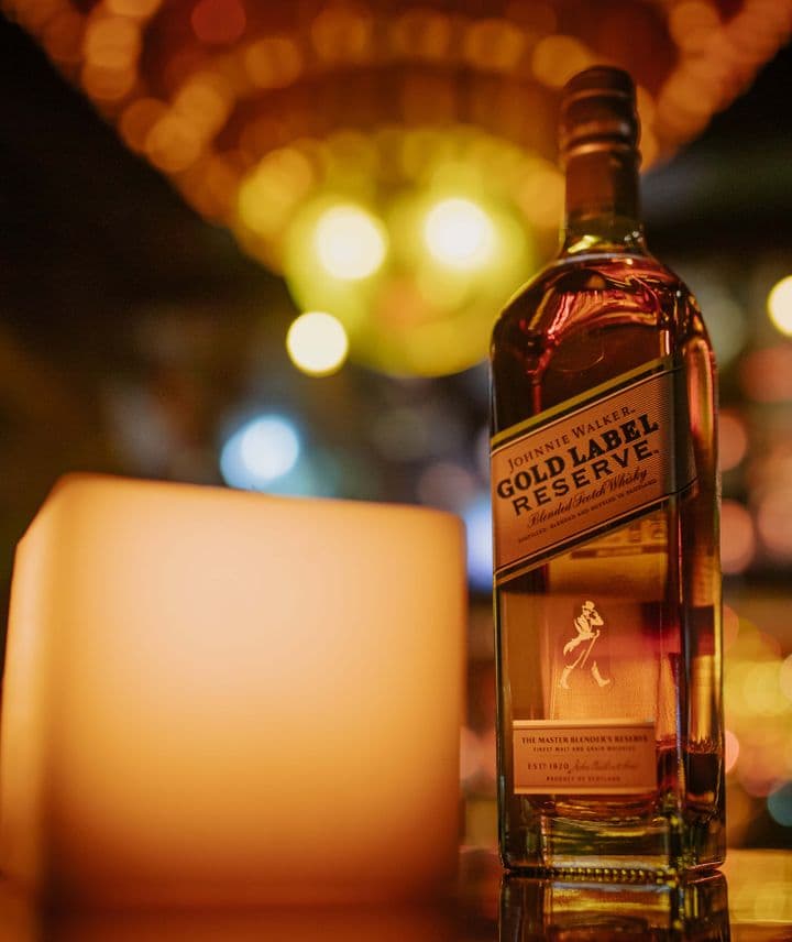 En la imagen se ve una botella de whisky Johnnie Walker Gold Label Reserve apoyado sobre una mesa y, a su lado, una vela. El ambiente es elegante y sofisticado.