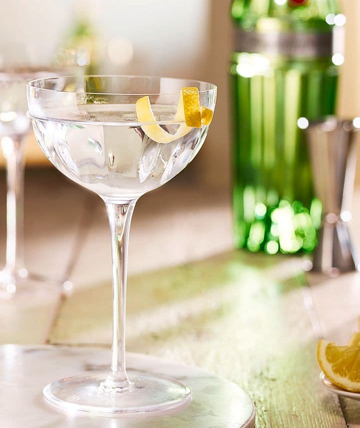 La imagen muestra dos copas con el cóctel Dry martini y, en el fondo, la botella de gin Tanqueray N°TEN y otros elementos de coctelería.