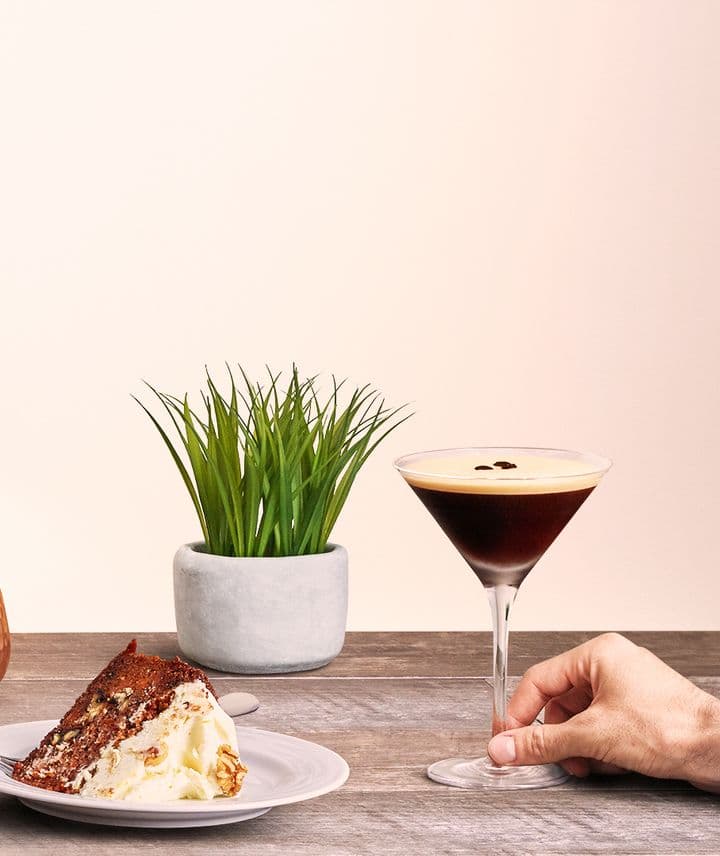 En la imagen se puede observar una copa que contiene el cóctel Espresso martini, acompañada por un plato con una porción de torta, y una maceta con una planta verde como decoración.