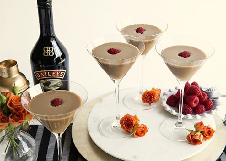 La imagen muestra 4 copas de martini con los cócteles French Dream, y alrededor se puede observar una botella de Baileys Original Irish Cream, flores naranjas y frambuesas.