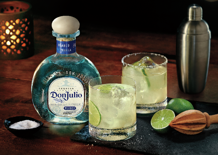 La imagen muestra una botella de tequila Don Julio Blanco junto a dos vasos con el cóctel Margarita y elementos de mixología alrededor.