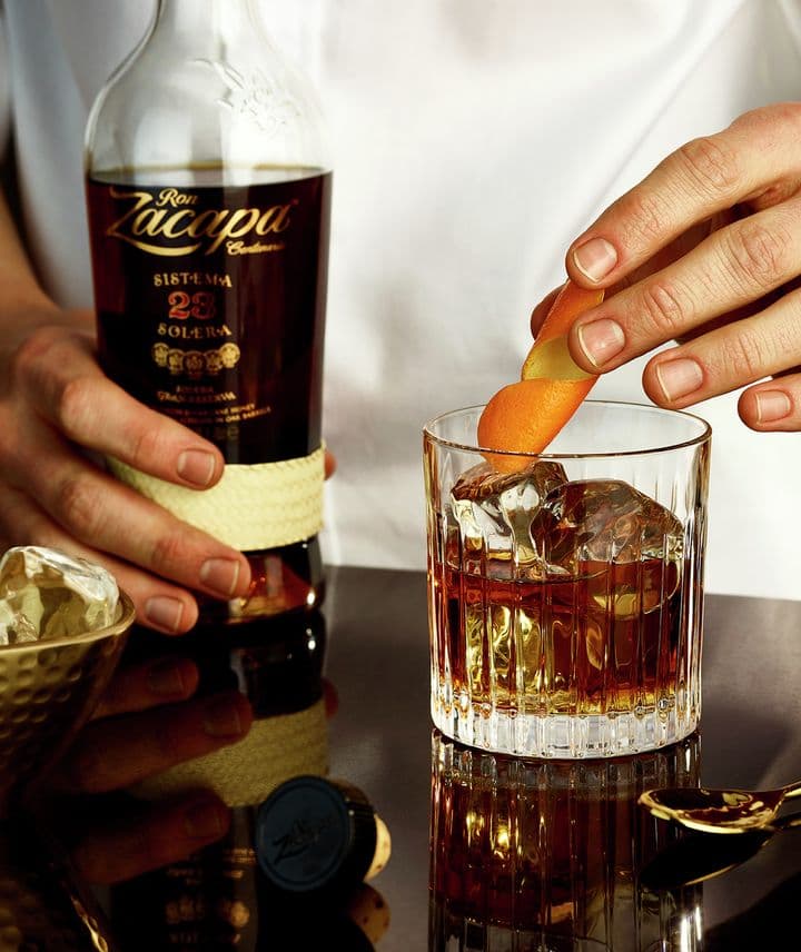 La imagen muestra a una persona agarrando una botella de ron Zacapa 23 y colocando una rodaja de naranja en un vaso que contiene el cóctel Old Fashioned.
