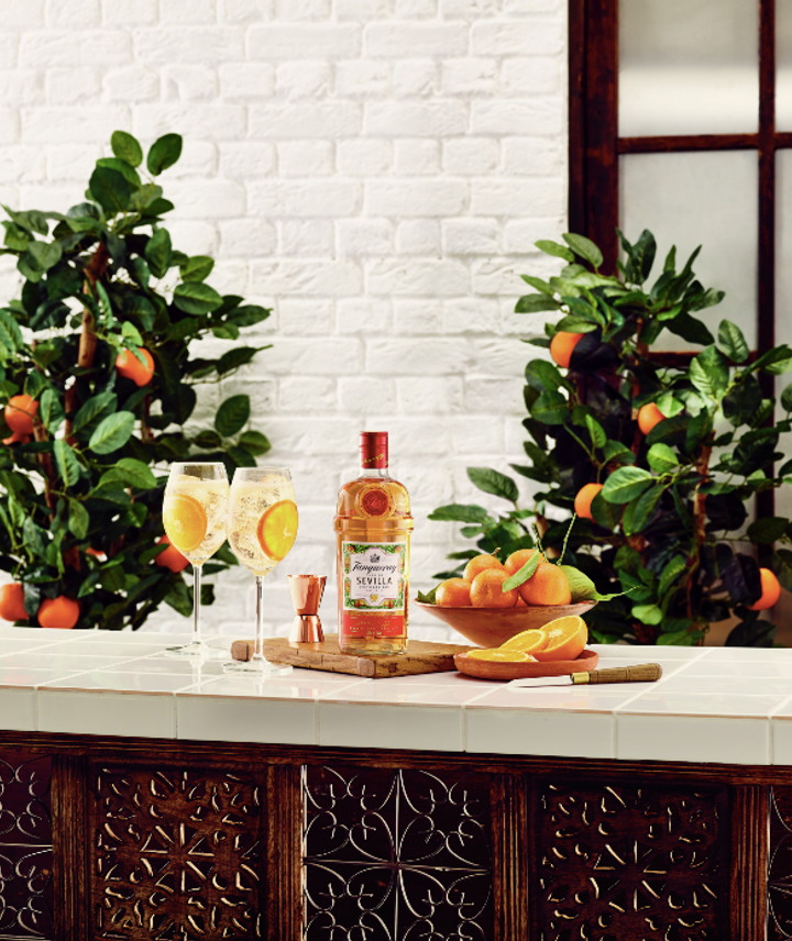 La imagen muestra una botella de gin Tanqueray Flor de Sevilla, dos copas con cócteles preparados con dicha bebida y elementos de coctelería sobre una mesada. De fondo, se observan dos árboles de naranja.