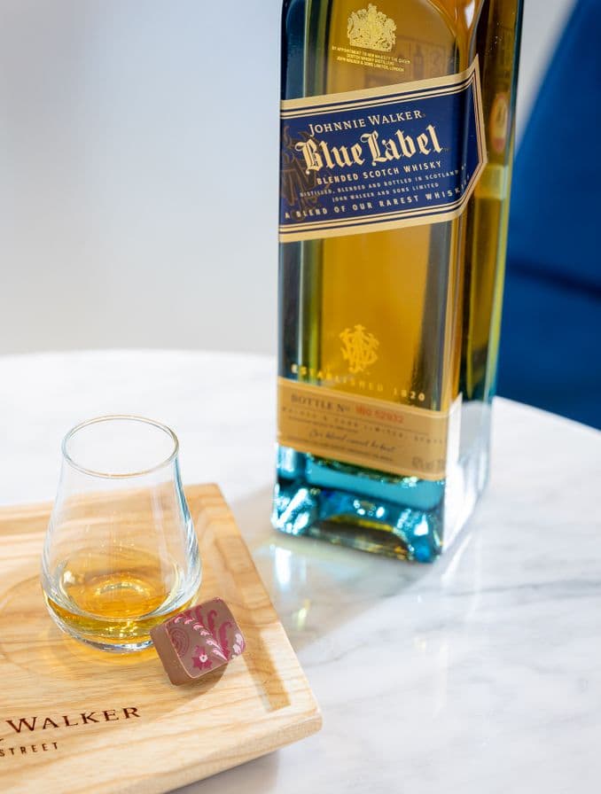 La imagen muestra una botella de whisky Johnnie Walker Blue Label junto a un vaso que contiene este destilado, con un chocolate ubicado a un lado.