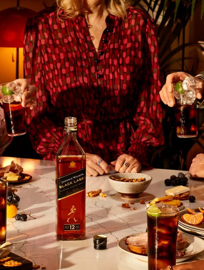 La imagen presenta una mesa con una variedad de platos, destacando en el centro una botella de whisky Johnnie Walker Black Label. Alrededor de la mesa, se observan personas, algunas de ellas sosteniendo vasos con cócteles.