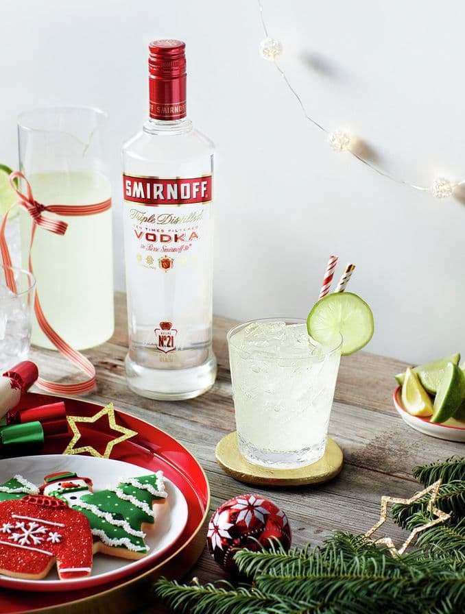 En la foto se puede apreciar una botella de Smirnoff No. 21 entre dos cócteles, cada uno en vasos de diferentes tamaños, todo ambientado en un entorno decorado con motivos navideños.