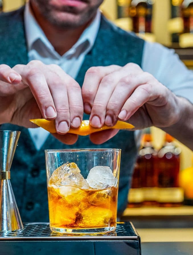 La imagen muestra un bartender en una barra preparando el cóctel Old-Fashioned.