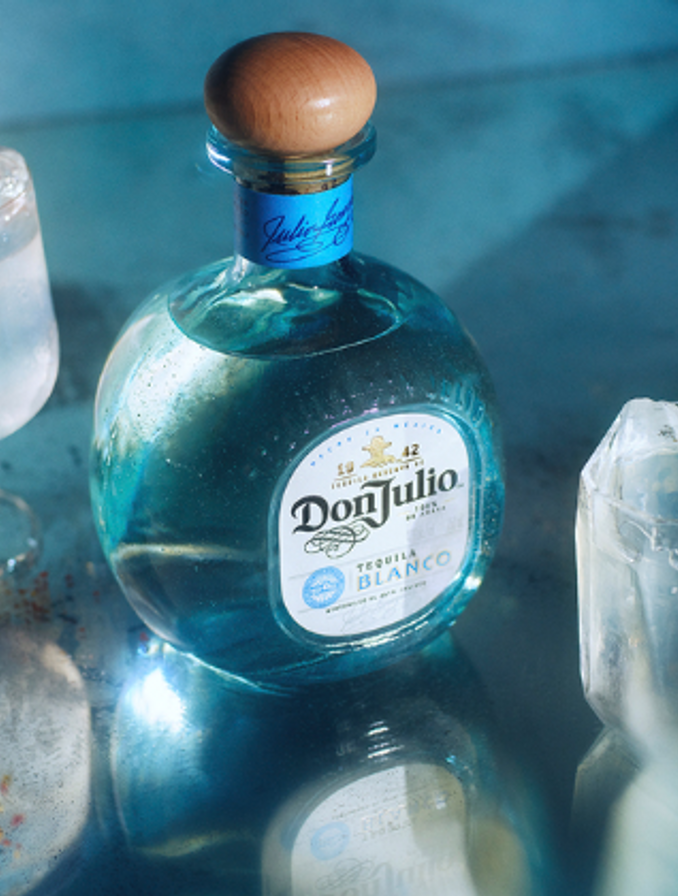 En la imagen se ve la botella de Don Julio Blanco sobre un fondo celeste. Lo rodean dos vasos con bebidas blancas y decoración.