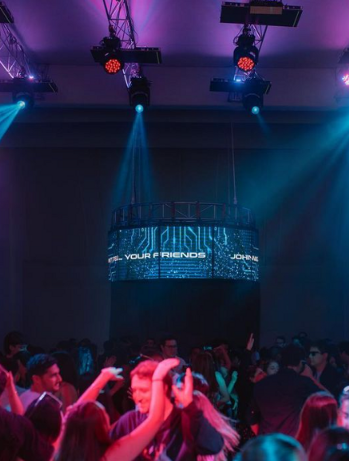 Imagen de discoteca, gente bailando , luces azules y violetas. De fondo la cabina del DJ a altura