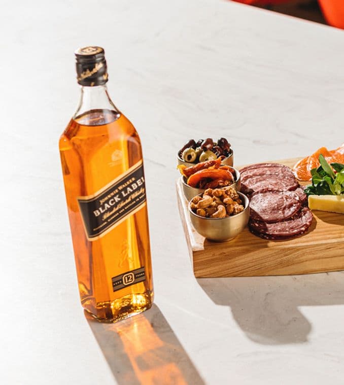 La imagen presenta una botella de whisky Johnnie Walker Black Label acompañada de una tabla con ingredientes salados y un vaso con un cóctel elaborado con dicho whisky.