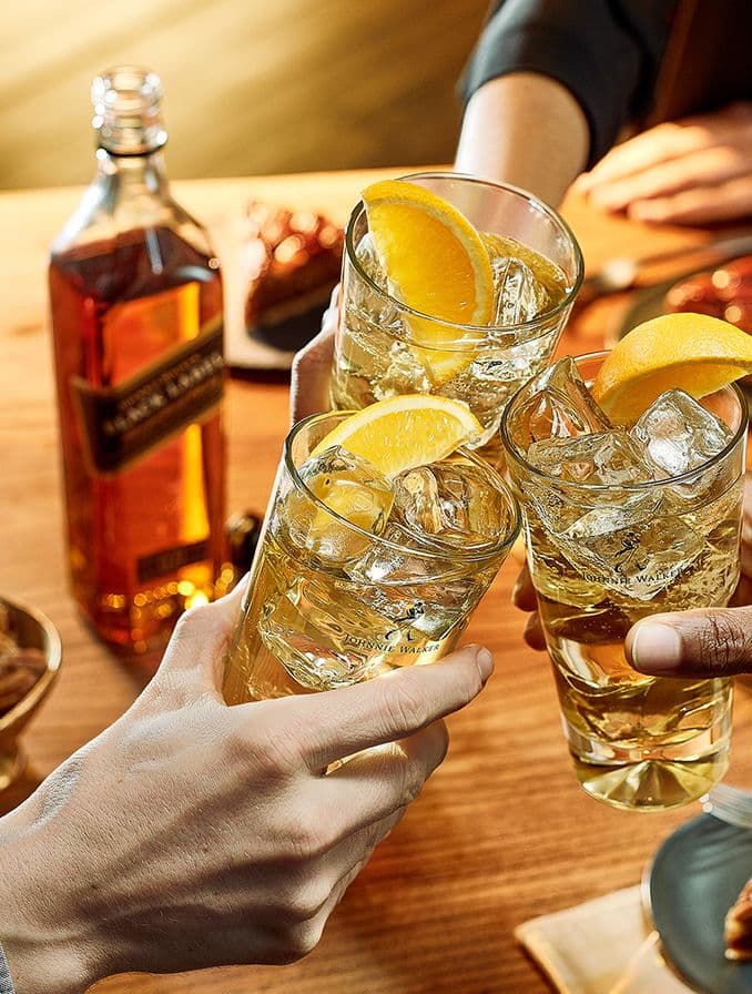 La imagen muestra tres manos sosteniendo cada una un vaso highball con whisky y limón, brindando. En segundo plano, se aprecia de manera difuminada una botella de whisky Johnnie Walker.