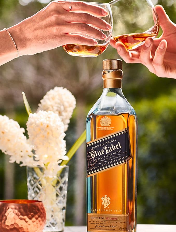 La imagen muestra una botella del whisky Johnnie Walker Blue Label, un brindis con dos vasos rellenos de dicho destilado y un florero con flores blancas.