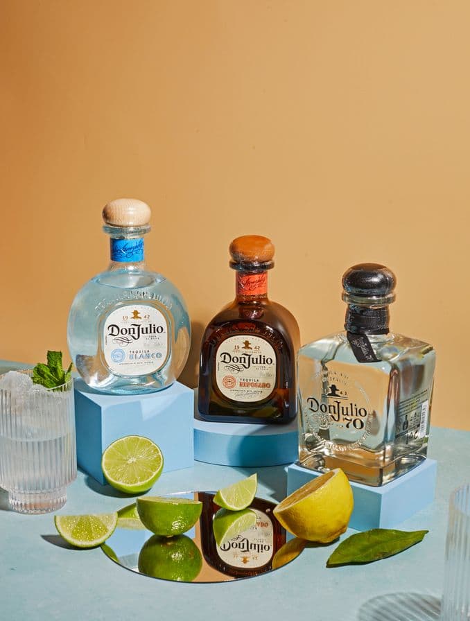 En la imagen se ven las botellas de Don Julio Reposado, Don Julio Blanco y Don Julio 70 sobre una tarima de color celeste. En el centro un espejo y limas y limones cortadas.