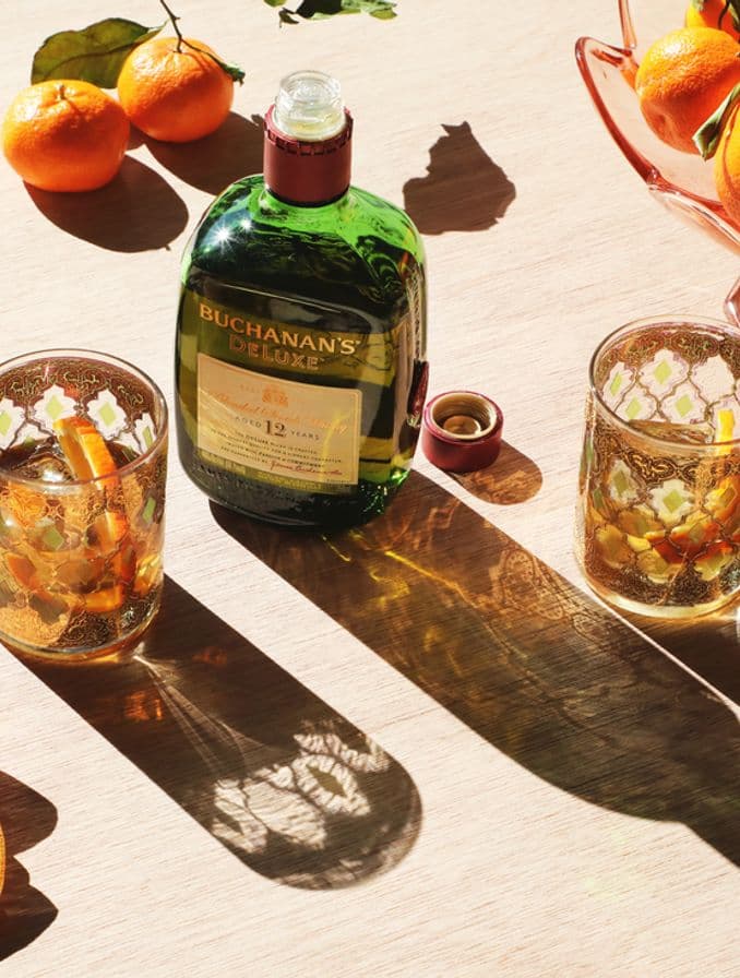 La imagen presenta una botella de whisky Buchanan's de 12 años junto a dos vasos con tragos preparados con dicho destilado, rodeados de un elemento de coctelería y naranjas.