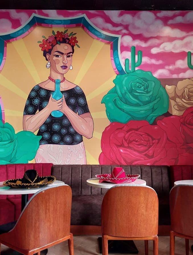En la imagen se ve un mural que decora el bar Rosa Mezcal, y las mesas a su alrededor. Decora en la mesa unos gorros mexicanos .