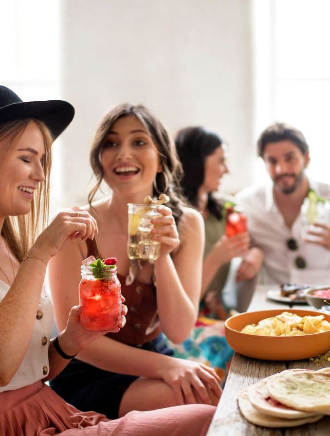 La imagen muestra cuatro personas, cada una con un cóctel en la mano, junto a una mesa con diversos platos de comida.